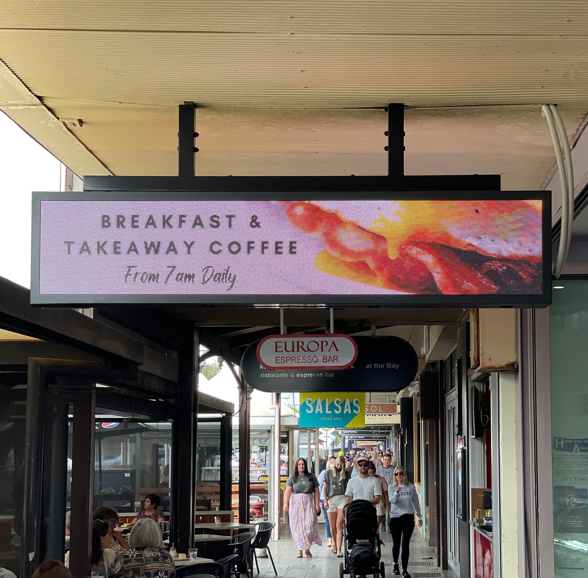 Hanging LED screen under a shopfront verandah, advertising breakfast and takeaway coffee.